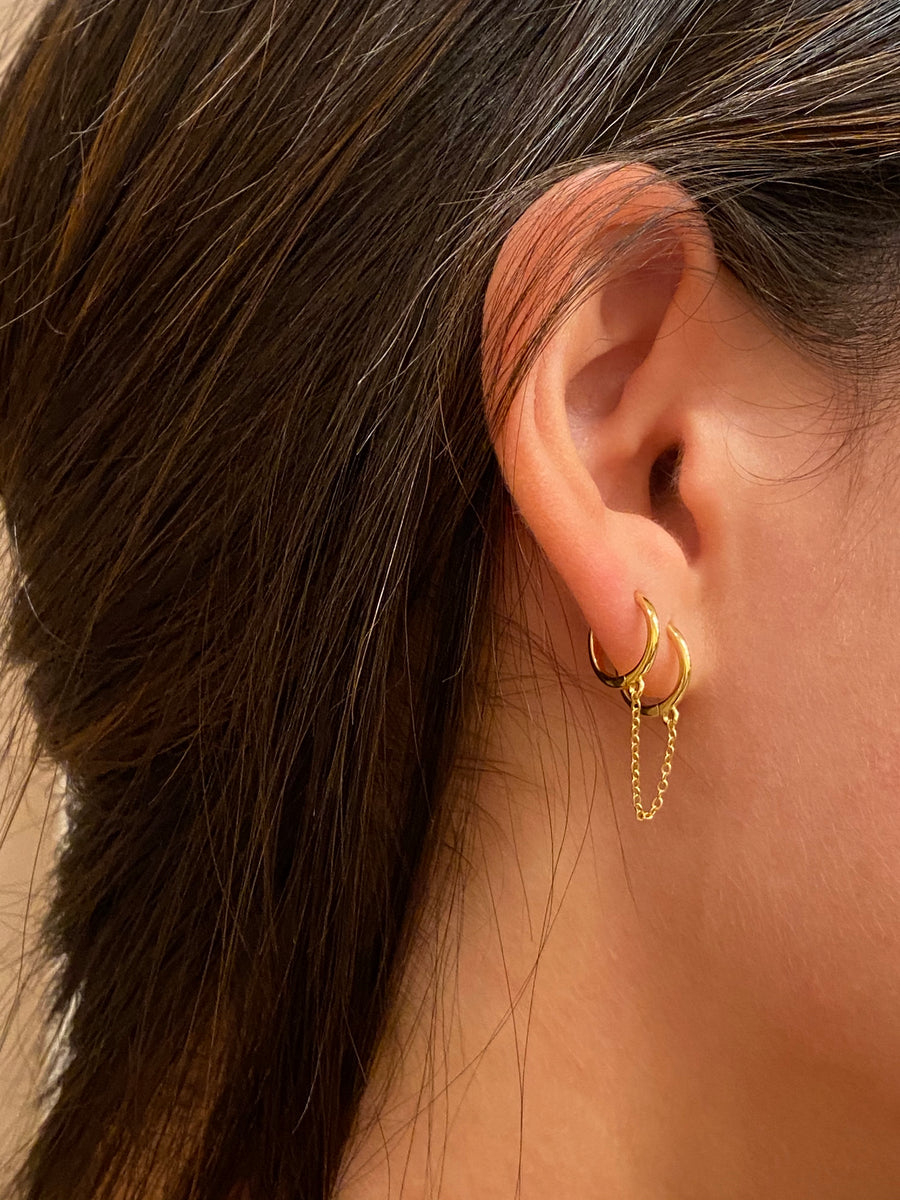 A close up of a woman's ear with a gold hoop in each hole, with a chain interlocking the two hoops.